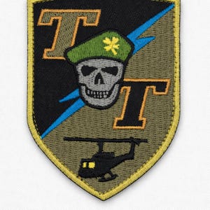 May include: Embroidered patch with a shield shape. The patch features a skull wearing a green beret with a yellow flower, a blue lightning bolt, and a helicopter. The letters "T" are in orange.