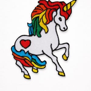 May include: Embroidered patch of a white unicorn with a rainbow mane and tail. The unicorn has a yellow horn and a red heart on its side. The patch is outlined in black and has a white background.