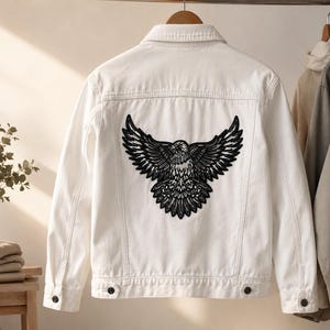 May include: A white denim jacket featuring a large, black, detailed graphic print of an eagle with its wings spread wide across the back panel. The jacket includes a classic collar, long sleeves with buttoned cuffs, and two chest pockets.