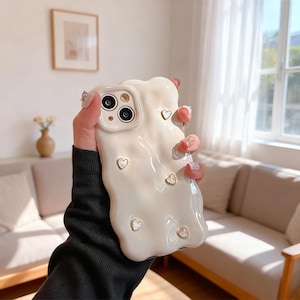 Puede incluir: Funda de teléfono blanca con un diseño texturizado y derretido y pequeños detalles de corazón dorado. La funda tiene un recorte para la cámara. La funda tiene una apariencia artística única.