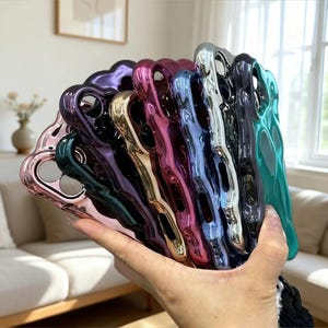 Puede incluir: Una colección de fundas de teléfono brillantes y coloridas en varios tonos, incluyendo rosa, verde, morado y plateado. Las fundas tienen una superficie lisa y reflectante y se mantienen juntas. Las fundas están diseñadas para proteger el teléfono.