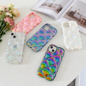 May include: A collection of iridescent phone cases in various colors, including pink, blue, and white. The cases feature a quilted design and are arranged on a white surface, with a bouquet of flowers in the background.