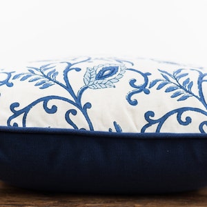 May include: A white pillow with a blue floral pattern and a navy blue border. The pillow is sitting on a wooden surface.