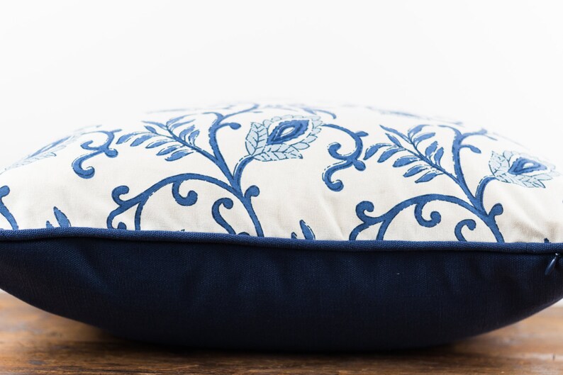 May include: A white pillow with a blue floral pattern and a navy blue border. The pillow is sitting on a wooden surface.