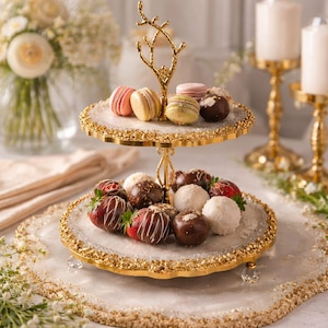 May include: A two-tiered gold dessert stand with a white and gold rim, displaying an array of treats. The top tier holds macarons, and the bottom tier features chocolate-covered strawberries and truffles. The stand is adorned with a gold, branch-like design.