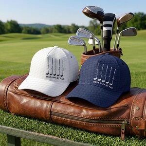 May include: A white and a navy blue baseball cap, each featuring a textured pattern and a graphic of five golf clubs with the text BEST PAPA BY PAR. The caps are positioned on a brown leather golf bag containing various metal golf clubs.