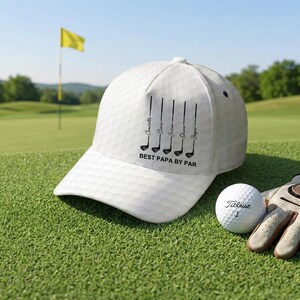 May include: A white baseball cap with a dimpled texture resembling a golf ball. The front features a black graphic of five golf clubs with the names Charlotte, Clara, Julian, Oliver, and Jasper printed vertically, and the text BEST PAPA BY PAR.