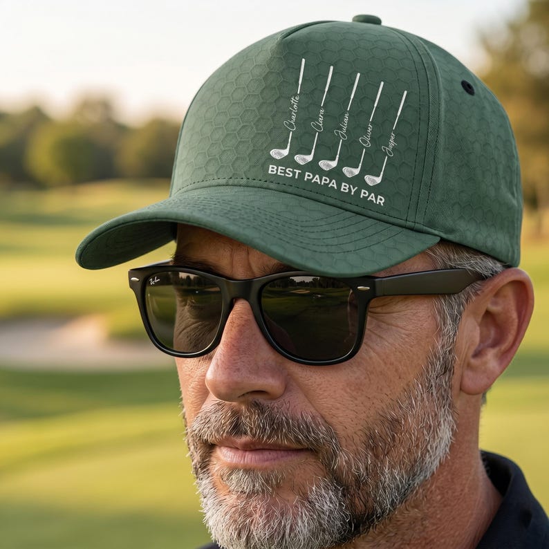 May include: A dark green baseball cap with a textured honeycomb pattern. The front features a white graphic of five golf clubs, each labeled with a name, and the text "BEST PAPA BY PAR" printed in white capital letters below the clubs.