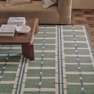 Könnte beinhalten: Rechteckiger Teppich mit grün-weißem geometrischen Muster und blauen Streifen. Im Hintergrund sind ein braunes Sofa und ein Couchtisch zu sehen, die eine gemütliche Atmosphäre schaffen.