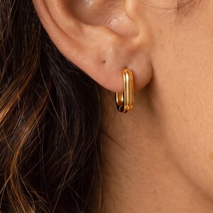 Puede incluir: Un pequeño pendiente de aro en tono dorado con forma rectangular alargada. El pendiente presenta un acabado metálico liso y está sujeto al lóbulo de la oreja, mostrando un diseño sencillo y limpio con bordes redondeados y una superficie brillante.