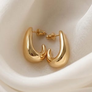 May include: A pair of gold-toned hoop earrings with a smooth, teardrop-shaped design. The earrings feature a polished, reflective surface and include standard post-back closures, showcasing a curved, open silhouette that tapers to a point.