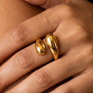 May include: A gold-toned open ring featuring two smooth, polished, teardrop-shaped elements of different sizes. The ring has a reflective metallic surface and a bypass design, with the two rounded ends positioned on the finger.