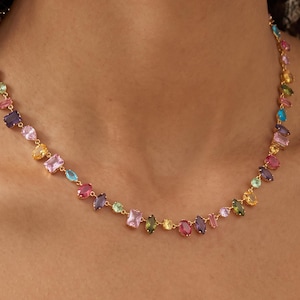 Peut inclure: Un collier à chaîne dorée orné d'une série de pierres précieuses multicolores de formes variées, notamment rectangulaires, ovales et en forme de poire. Les pierres présentent une palette vibrante de tons roses, bleus, jaunes, violets et verts.