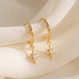 May include: A pair of gold hoop earrings featuring a small star-shaped charm on each hoop. Each charm is accented with a single clear, round-cut gemstone at its center. The hoops also feature a small, fixed star design on the upper band.
