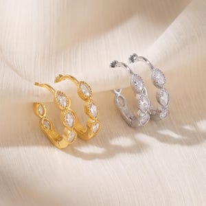 May include: A pair of gold-toned hoop earrings and a pair of silver-toned hoop earrings, each featuring a series of marquise-cut clear stones set along the outer edge of the circular band, creating a reflective and detailed jewelry design.