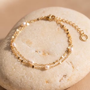 Puede incluir: Una delicada pulsera de cadena dorada que presenta dos hilos finos y brillantes unidos entre sí. La pulsera está adornada con pequeñas perlas blancas redondas espaciadas uniformemente a lo largo de la cadena e incluye un pequeño cierre circular.