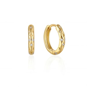 May include: A pair of gold-colored hoop earrings. Each earring features a starburst pattern with small, clear gemstones. The earrings are displayed against a white background, highlighting their design and color.