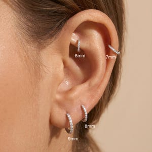 May include: Close-up of an ear adorned with four different sizes of small, silver hoop earrings. The earrings are encrusted with small, sparkling stones. Sizes are 6mm, 7mm, 8mm, and 9mm.