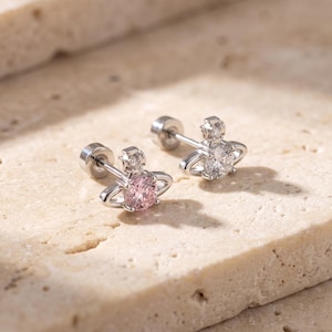 May include: A pair of silver-toned stud earrings featuring a planet-shaped design with a central round gemstone. One earring contains a light pink stone, while the other features a clear white stone. Each earring has a small cross detail on top.