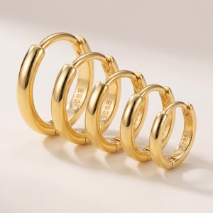 May include: A collection of four gold-coloured hoop earrings in different sizes. The earrings are displayed in a line, with the largest hoop on the left, decreasing in size to the right. Each earring has a smooth, polished finish and a simple, classic design.