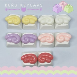 Pode incluir: Uma coleção de keycaps personalizadas em várias cores pastel, incluindo amarelo, branco, roxo, rosa e vermelho. Cada keycap apresenta um design único em forma de asa. O texto "BERU KEYCAPS" é exibido no topo.