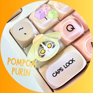 Pode incluir: Uma tecla de teclado personalizada com um personagem de desenho animado em uma roupa amarela. A tecla est&aacute; em um teclado branco com outras teclas em tons de branco e rosa. O texto "POMPOM PURIN" e "CAPS LOCK" s&atilde;o vis&iacute;veis.