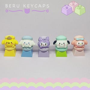 以下が含まれることがあります： 様々なキャラクターがデザインされた、カラフルなキーキャップが5つ並んでいます。各キーキャップは異なる色とデザインで、正方形のベースに取り付けられています。「BERU KEYCAPS」の文字が上部に表示されています。