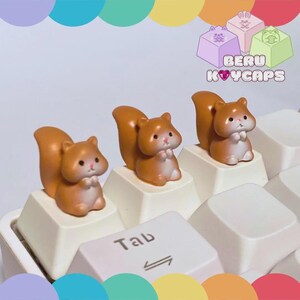 May include: Three brown squirrel keycaps on a white keyboard. The keycaps are shaped like small squirrels with fluffy tails and are positioned on the keys. The image also includes the text "BERU KOYCAPS".