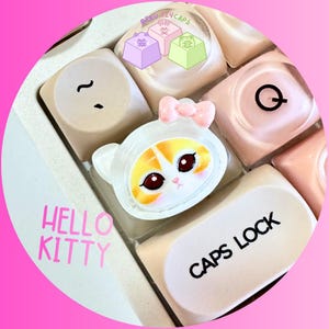 Pode incluir: Close-up de teclas personalizadas, incluindo um design Hello Kitty com um la&ccedil;o rosa. As teclas s&atilde;o bege e rosa, com um design de gato branco e laranja. Os textos "HELLO KITTY" e "CAPS LOCK" s&atilde;o vis&iacute;veis.