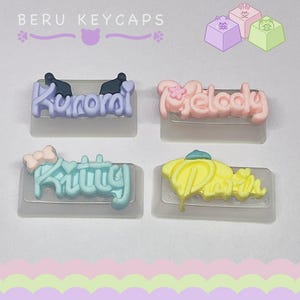 Puede incluir: Cuatro teclas de teclado coloridas con los nombres de los personajes: Kuromi, Melody, Kitty y Pompompurin. Las teclas son de colores pastel, cada nombre está escrito en una fuente diferente. El texto "BERU KEYCAPS" también es visible en la parte superior.