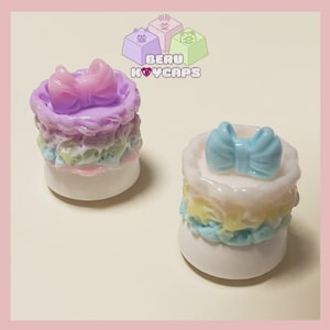 May include: Two miniature cake-shaped keycaps with bow accents. One keycap features a purple bow and frosting, the other has a blue bow. The keycaps have layers of pastel colors and are set against a light pink background. The text "BERU KEYCAPS" is visible.