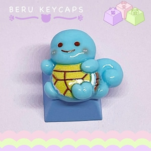 May include: A light blue keycap featuring a cartoon turtle character with a yellow shell and brown accents. The keycap is mounted on a blue base. The text "BERU KEYCAPS" is above the keycap.
