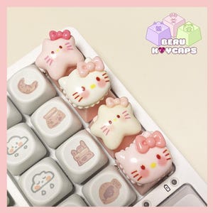 May include: A close-up of a white keyboard with custom keycaps. The keycaps feature cute, cartoon-style designs of a cat and star shapes in pink and white, with bows. The keycaps are on a white keyboard. The logo "BERU KEYCAPS" is in the upper right corner.