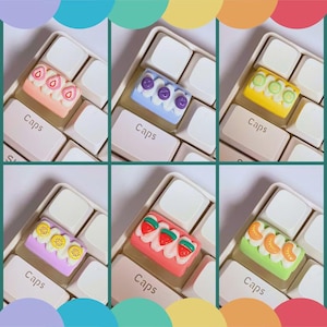 May include: Six colorful artisan keycaps, each designed to resemble a miniature cake with fruit toppings. The keycaps are set on a white keyboard, with the word "Caps" visible on the keys. The fruit toppings include strawberries, blueberries, kiwi, and oranges.