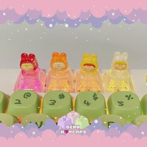May include: A close-up of a keyboard with custom keycaps. The keycaps are light green with black symbols. Four translucent keycap figures in red, orange, yellow, and white are on top. The brand name "BERU KOYCAPS" is visible.