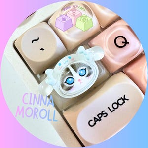 Pode incluir: Close-up de teclas de teclado personalizadas. Uma tecla apresenta um design de gato branco com olhos azuis e um la&ccedil;o azul. Outras teclas t&ecirc;m um tom rosa claro. O texto "CINNA MOROLL" &eacute; vis&iacute;vel. As teclas fazem parte de um teclado.