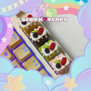 May include: A collection of colorful, cake-shaped keycaps with white frosting, berries, and the text "BERU KEYCAPS." The keycaps are arranged on a keyboard, with additional keycaps in the background. The image has a pastel color scheme.