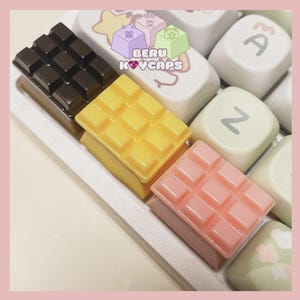 May include: Close-up of a keyboard with custom keycaps shaped like chocolate bars in brown, yellow, and pink. The keycaps are on a white keyboard with other keycaps that have cartoon designs and the text "BERU KEYCAPS".
