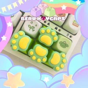 Puede incluir: Primer plano de un teclado con teclas personalizadas. Las teclas presentan un diseño de huella de pata verde con almohadillas amarillas. Otras teclas tienen ilustraciones de un gato, una rana y otros diseños. El texto "BERU KEYCAPS" es visible.