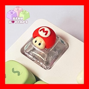 May include: A red and white mushroom-shaped keycap with a white "M" symbol, set on a clear, square base. The keycap is part of a keyboard, with other colorful keycaps visible. The text "BERU KEYCAPS" is in the upper left corner.