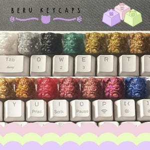 May include: A close-up of a keyboard with custom keycaps. The keycaps are various colors, including gold, red, and blue, and have a textured, sculpted design. The keyboard keys are white, and the image includes the text "BERU KEYCAPS".