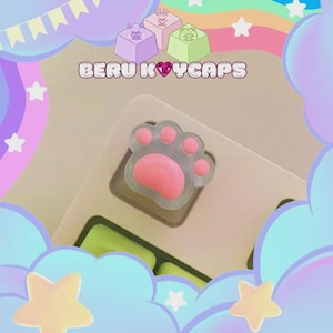 Könnte beinhalten: Ein klarer Keycap in Form einer Katzenpfote mit einem rosa Pfotenballen und rosa Zehenballen. Der Keycap befindet sich auf einer Tastatur mit grünen Tasten. Der Text "BERU KEYCAPS" steht über dem Keycap. Der Hintergrund hat Pastellfarben und Sterne.