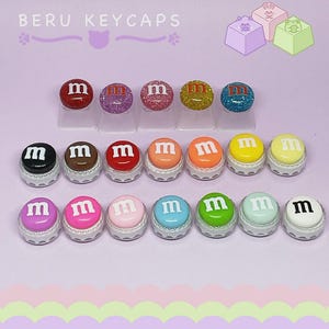 May include: A collection of colorful keycaps with the letter "m" on them. The keycaps are in various colors, including red, pink, yellow, and blue. The text "BERU KEYCAPS" is at the top of the image.