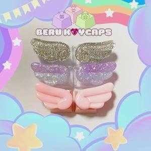 May include: A collection of colorful keycaps with wing designs. The keycaps are in silver glitter, purple and white, and pink. The text "BERU KEYCAPS" is displayed above the keycaps. The background is a pastel rainbow with stars and clouds.