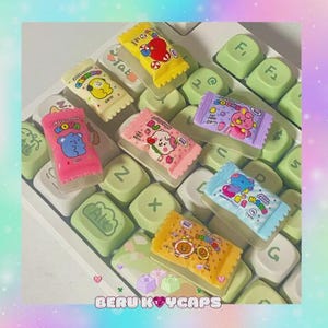 Könnte beinhalten: Eine Sammlung von bunten, bonbonförmigen Tastenkappen mit Cartoon-Charakter-Designs, die auf einer weißen Tastatur mit grünen Tasten liegen. Die Tastenkappen haben verschiedene Farben wie Rosa, Gelb, Blau und Lila, mit Text wie "ROYA", "COOKY" und "SHOO".