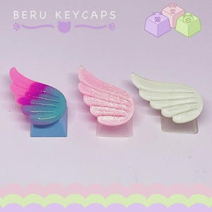 Pode incluir: Três keycaps em forma de asa em rosa, azul e branco. As keycaps são montadas em bases de plástico transparente. O texto "BERU KEYCAPS" é visível na parte superior da imagem. Fundo roxo claro.