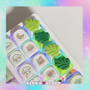 May include: A custom keyboard with white keycaps featuring food-themed designs and green leaf-shaped keycaps. The keyboard has a colorful, illuminated base and the text "BERU KEYCAPS" is visible.