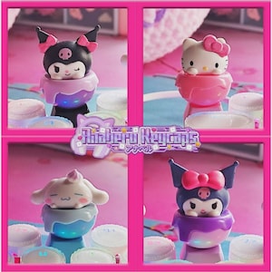 May include: Four colorful keycaps featuring popular characters. The keycaps include Hello Kitty, Kuromi, and Cinnamoroll. The keycaps are set against a pink background with a floral pattern. The text "Amaberri Keycaps" is also visible.