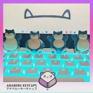 May include: Four Snorlax keycaps sit on a backlit keyboard. The keycaps are blue and beige, with a cartoonish design. The keyboard has white keys and a light blue glow. The image also includes a graphic of a Snorlax's ears and the text "ANABERU KEYCAPS".