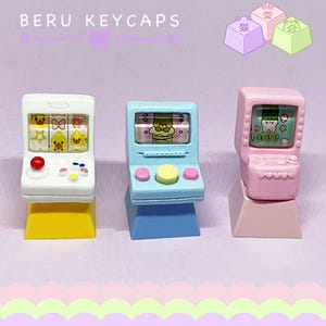 Könnte beinhalten: Drei farbenfrohe Tastenkappen in Form von Miniatur-Spielautomaten. Die Tastenkappen sind weiß, blau und rosa, mit gelben, roten und rosa Knöpfen. Die Bildschirme zeigen Cartoon-Grafiken. Der Text "BERU KEYCAPS" steht oben.